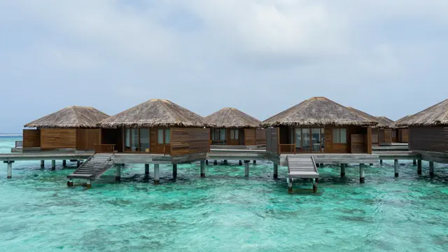 Row of wooden overwater bungalows with thatched roofs standing above a turquoise lagoon under a pale cloudy sky