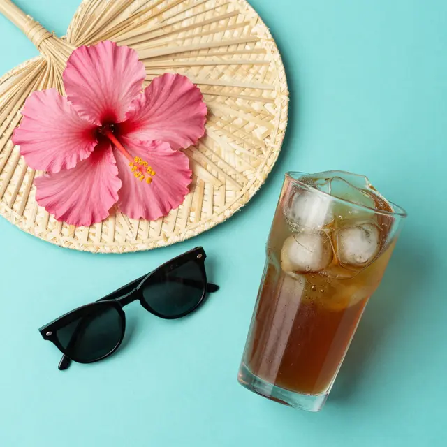 Top-down flat lay of a straw fan, pink hibiscus, sunglasses and iced tea on a turquoise background