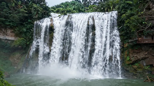 A wide cascading waterfall flows into a green pool surrounded by dense jungle vegetation