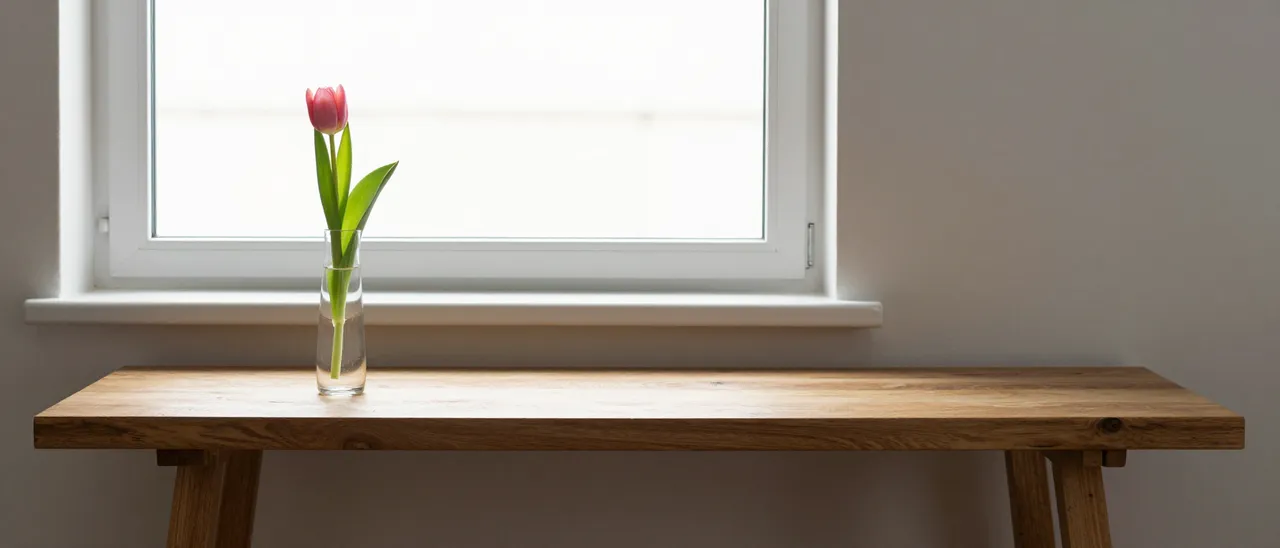 A single pink tulip in a glass bud vase sits on a long oak console table by a bright window in soft morning light.