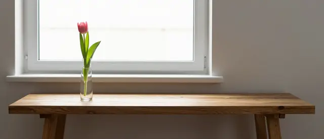 A single pink tulip in a glass bud vase sits on a long oak console table by a bright window in soft morning light.