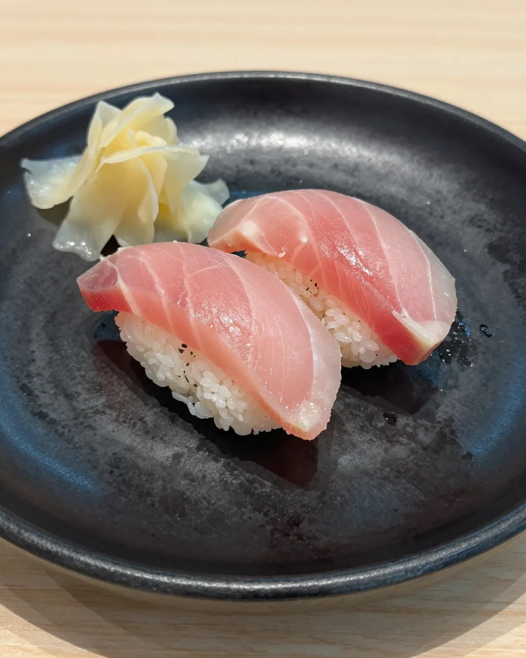 Two pieces of tuna nigiri and pickled ginger rest on a matte black ceramic plate atop a light wooden table.