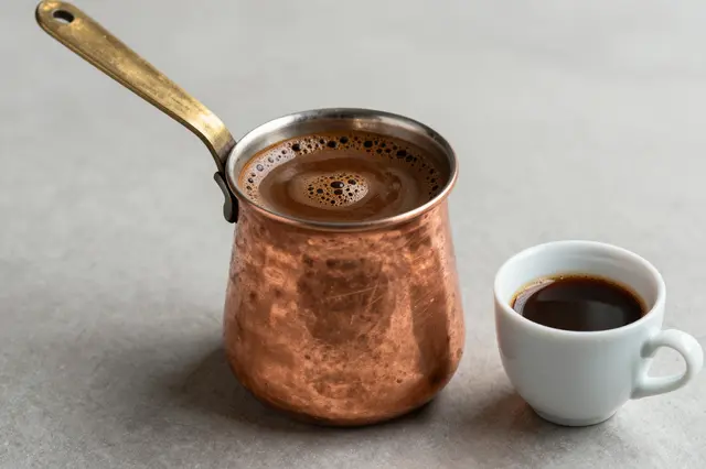 Hammered copper cezve of frothy Turkish coffee sits next to a small white filled coffee cup on a smooth gray countertop