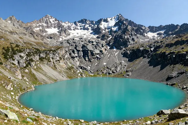 Vibrant turquoise alpine lake sits below jagged snow-dusted Swiss mountain peaks under a clear blue sky