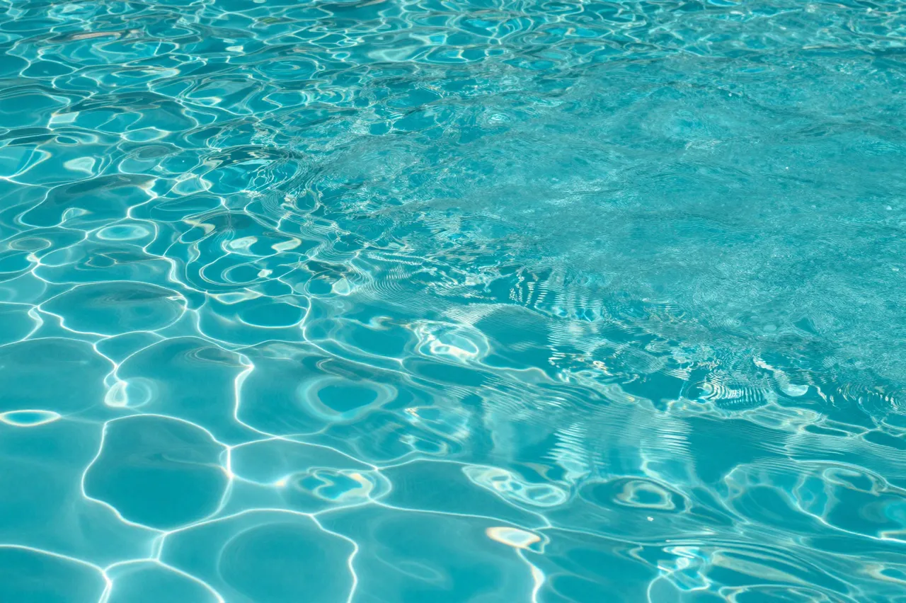 Top view of rippled bright turquoise swimming pool water with glowing sun reflection patterns across the full frame.