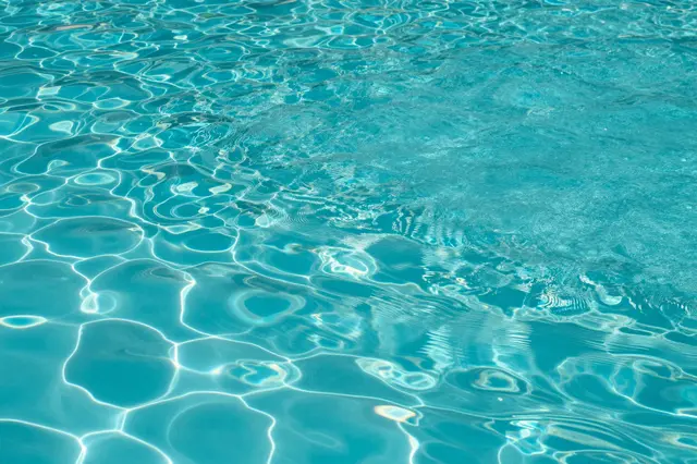 Top view of rippled bright turquoise swimming pool water with glowing sun reflection patterns across the full frame.