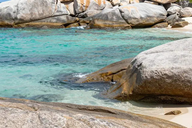 Clear turquoise coastal water laps against smooth weathered granite boulders on a sunny beach