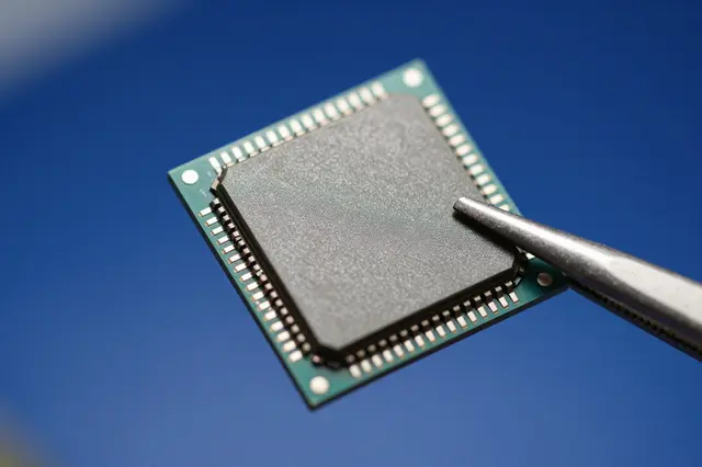 A macro close-up of a blank silicon chip held by metal tweezers against a soft blue background