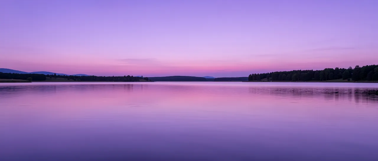 A smooth calm lake reflects lavender and lilac twilight sky above tree lined shores