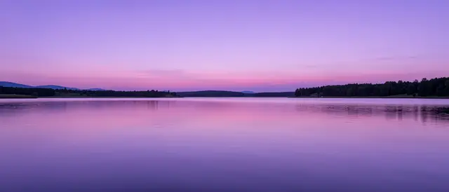 A smooth calm lake reflects lavender and lilac twilight sky above tree lined shores
