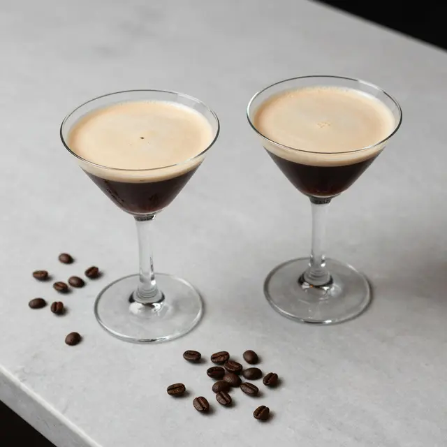 Top view of two foamy espresso martinis with scattered coffee beans on a light marble bar top