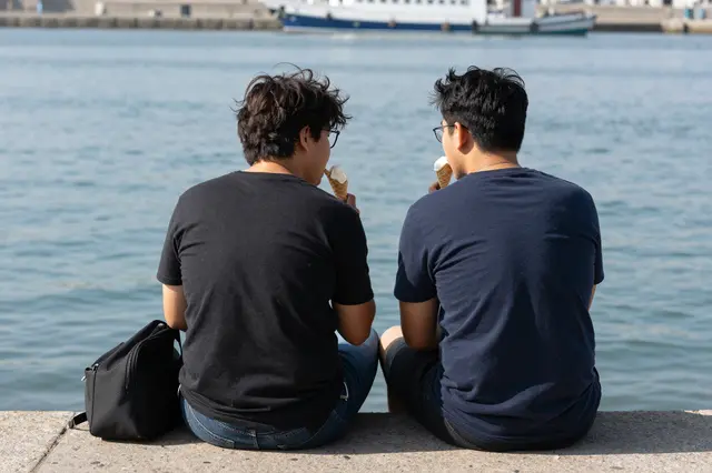 Two young men sit on a concrete harbor wall from behind, eating ice cream cones overlooking calm water with distant boats.