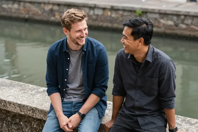 Two men in their thirties sit on a stone canal wall laughing together as they look at each other