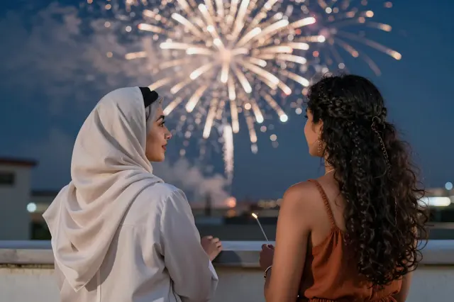 Two women stand on a rooftop at dusk watching bright golden fireworks burst in the night sky.