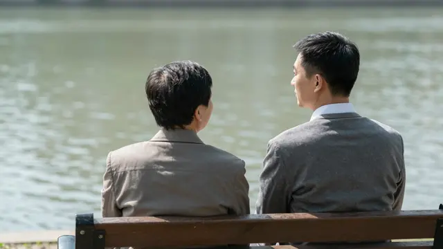 Two East Asian men viewed from behind sit on a park bench looking out over calm river water