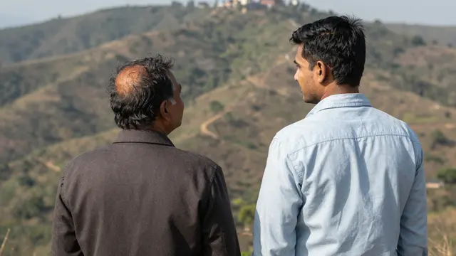 Back view of a younger South Asian man and an older man standing on a hilltop looking out at rolling hills