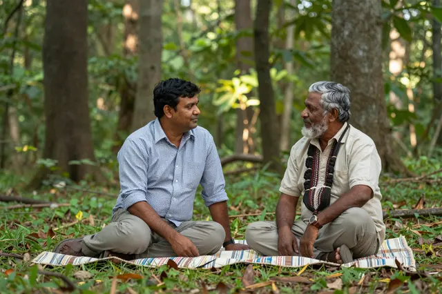 Two older men sit cross-legged on a striped blanket in a sun-dappled forest clearing, facing each other.