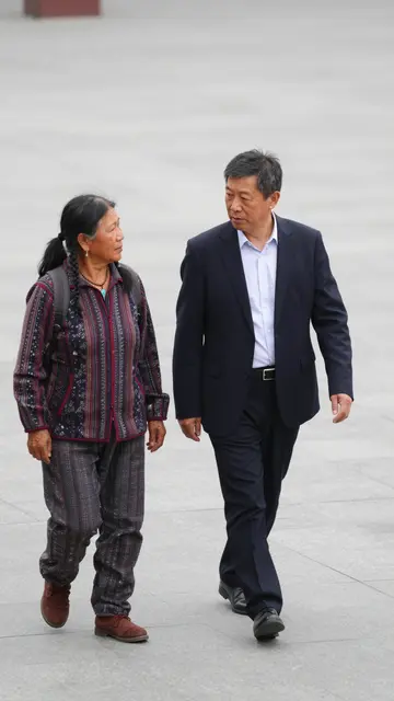 An older Indigenous woman and an older East Asian man walk side by side conversing across a paved stone plaza.