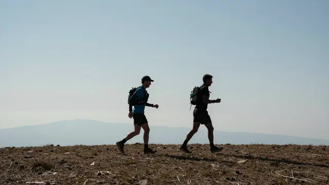 Two adult trail runners travel side by side across a dry rocky high plateau under a hazy pale sky with distant mountains.
