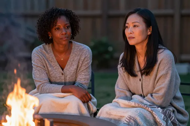 Two women sit covered with blankets by a glowing backyard fire pit at dusk