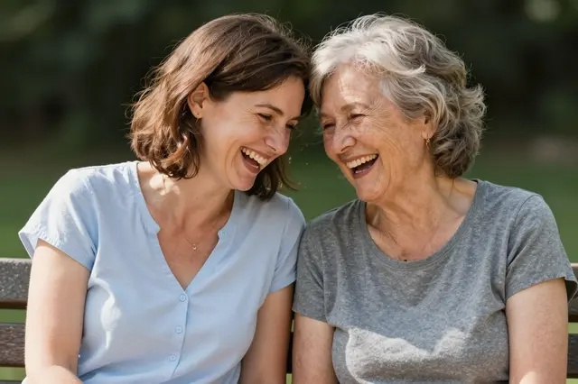 A middle aged woman and an older woman sit close together laughing on a sunny park bench