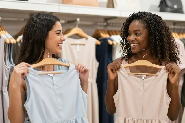 Two smiling women laugh together while holding plain dresses on hangers in a clothing boutique
