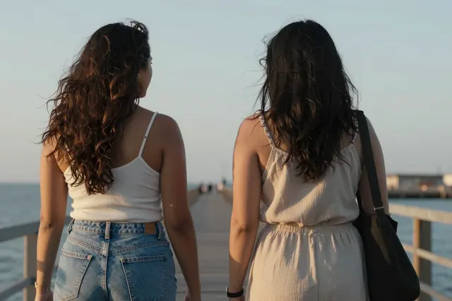 Two women viewed from behind walk side by side along a wooden pier at golden hour by the sea
