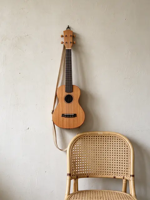 Wooden ukulele hangs on a pale plaster wall above a woven rattan chair in soft daylight