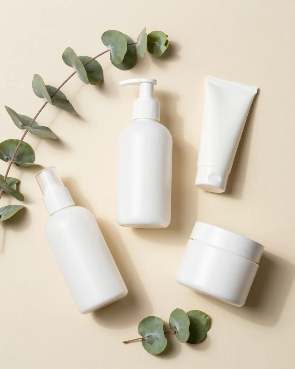 Flat lay of blank white skincare containers arranged with eucalyptus sprigs on a soft beige background