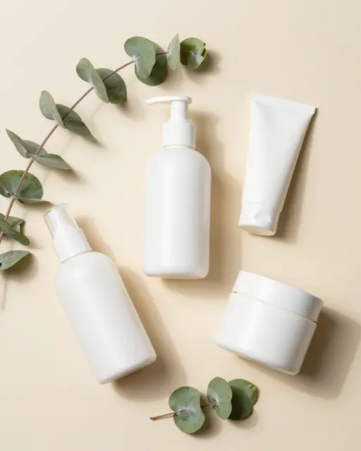Flat lay of blank white skincare containers arranged with eucalyptus sprigs on a soft beige background