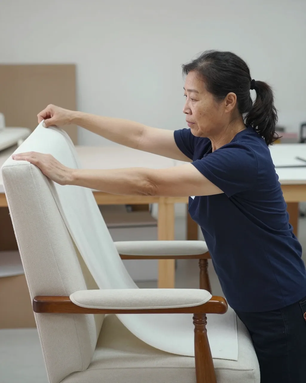 Side view of an older East Asian woman stretching fabric over an armchair frame in a bright workshop.