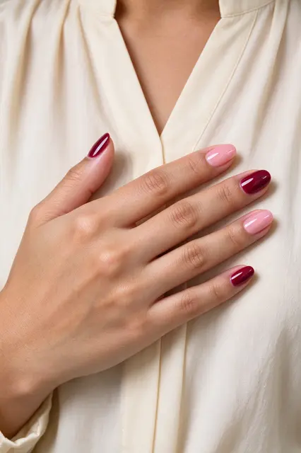 A woman's hand with alternating burgundy and blush oval nail art rests on a cream blouse