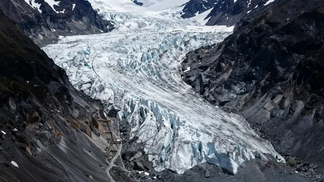 A large crevassed glacier flows down a steep rocky valley between dark jagged mountain slopes