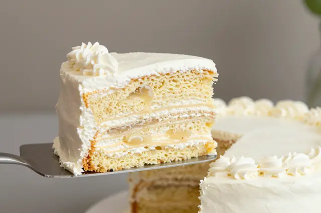 A cake server lifts a frosted vanilla layer slice away from the remaining whole cake.