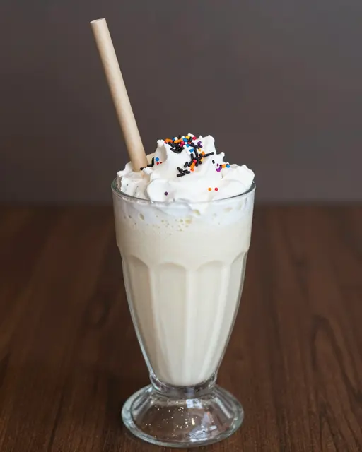 A vanilla milkshake topped with whipped cream, colorful sprinkles, and a paper straw sits in a glass on a wooden table.