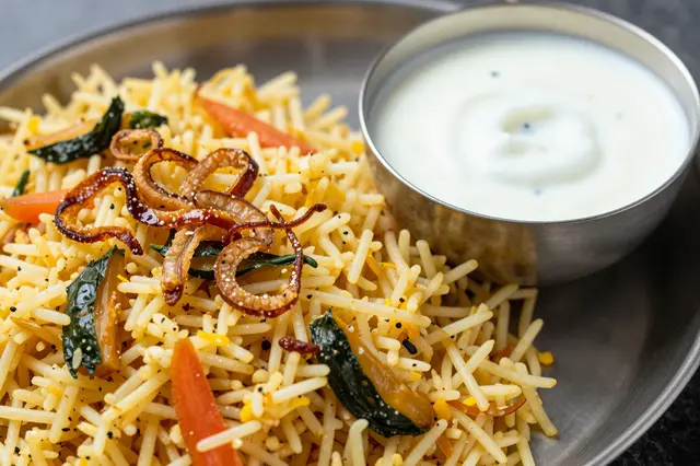 Close-up of vegetable biryani topped with fried onions, served with a metal bowl of creamy raita on a thali plate.