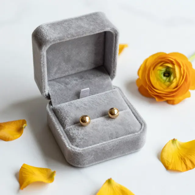 Gray velvet jewelry box holding gold stud earrings sits with yellow ranunculus petals on white marble