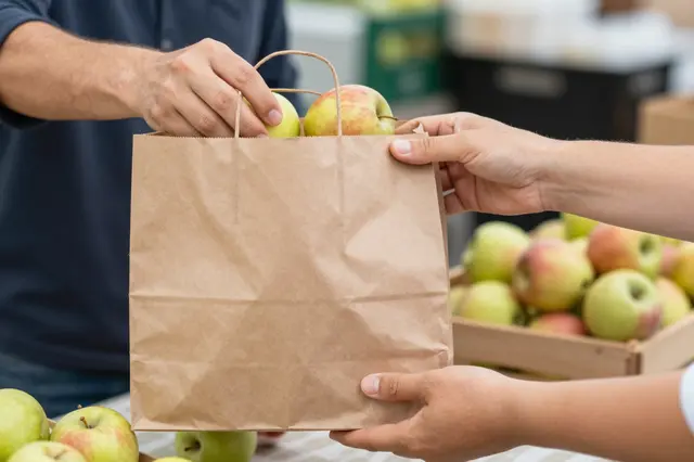 Vendor passes a brown paper bag of apples to a customer over a farmers market stall table