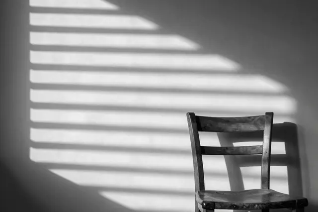 Black and white view of an empty wooden chair below sharp striped venetian blind shadows cast on a plain plaster wall
