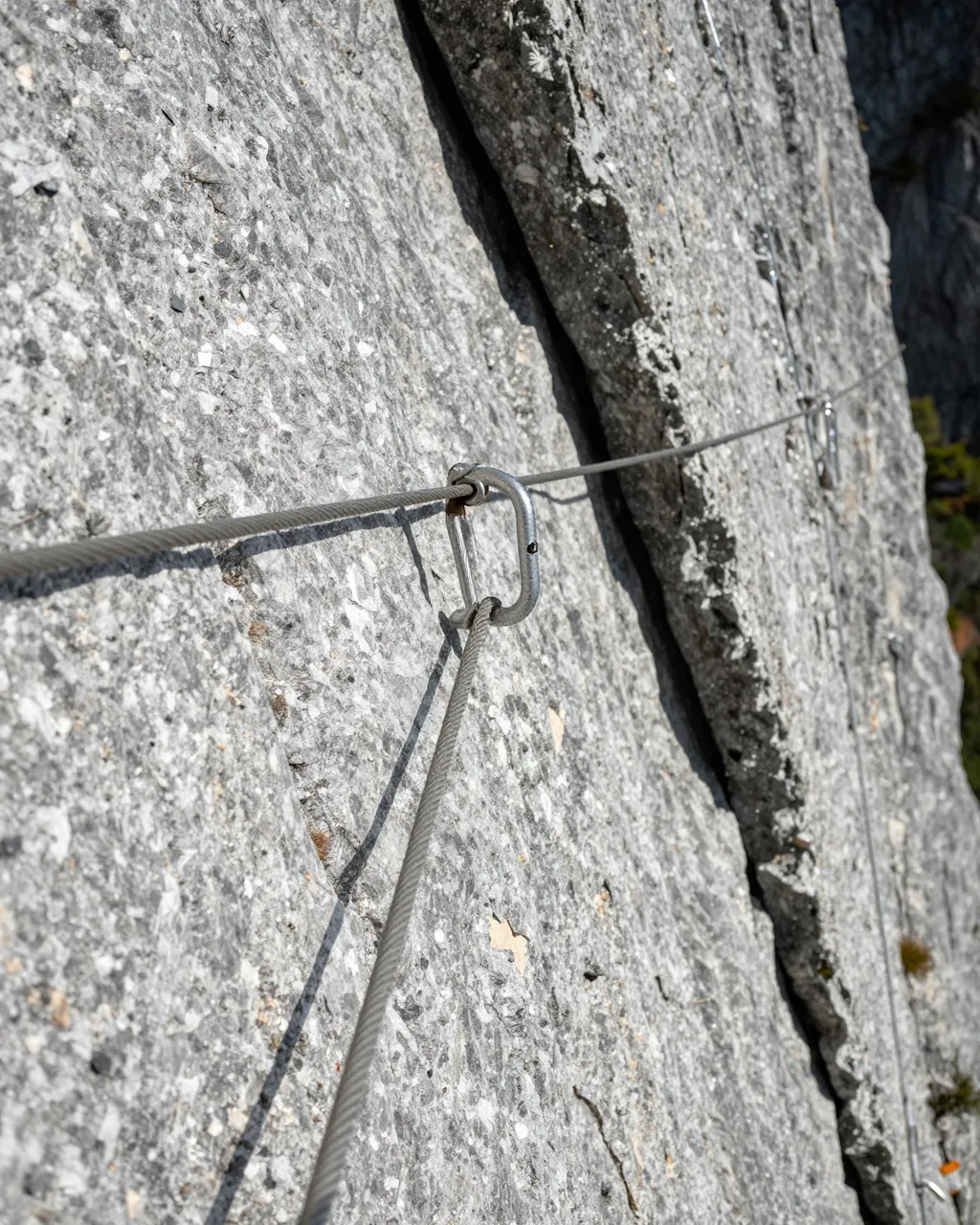 Close-up view of a via ferrata steel cable and carabiner mounted to a sunlit gray granite cliff face