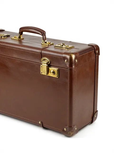 A side-angle view of a vintage brown leather suitcase with brass hardware on a plain white studio background