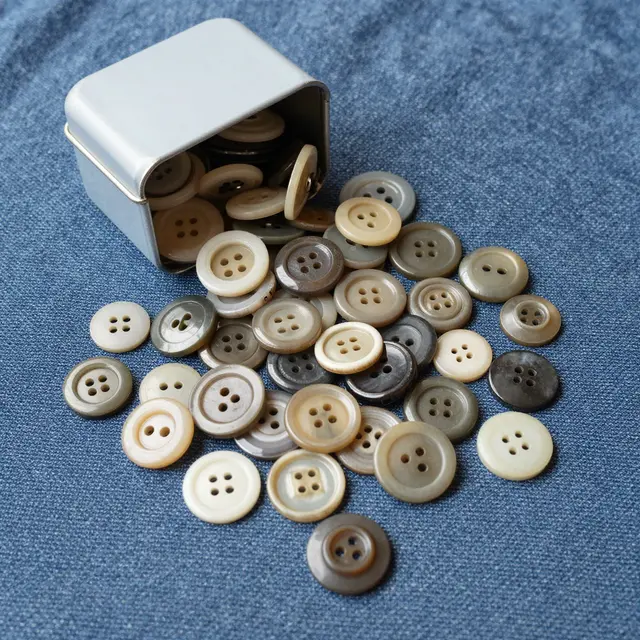 A small plain tin spills assorted neutral tone vintage buttons onto blue denim fabric
