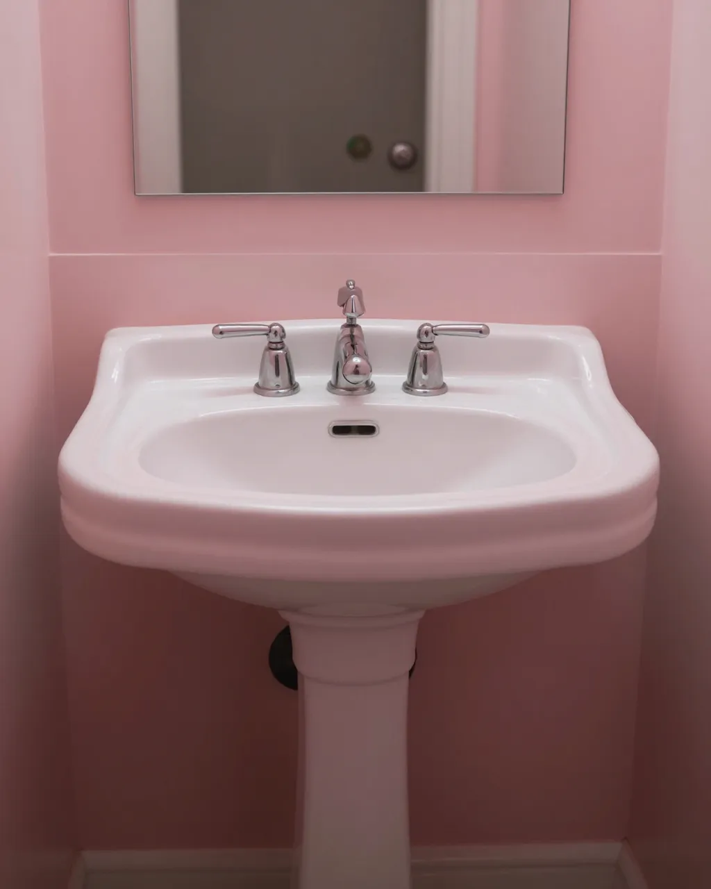 White vintage pedestal sink with chrome faucets sits against soft pink bathroom walls below a wall mirror