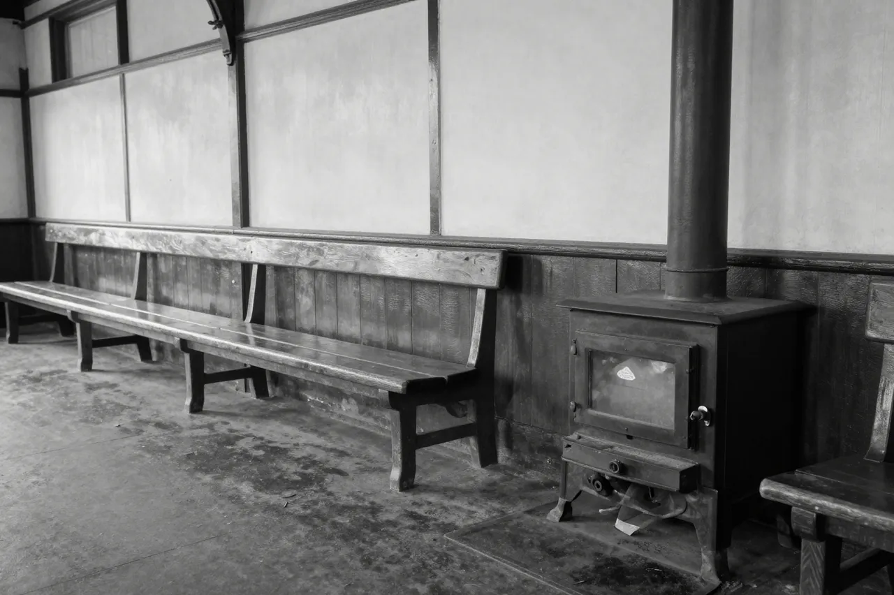 Black and white view of an empty old railway waiting room with long wooden benches and a cast iron stove