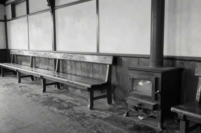 Black and white view of an empty old railway waiting room with long wooden benches and a cast iron stove