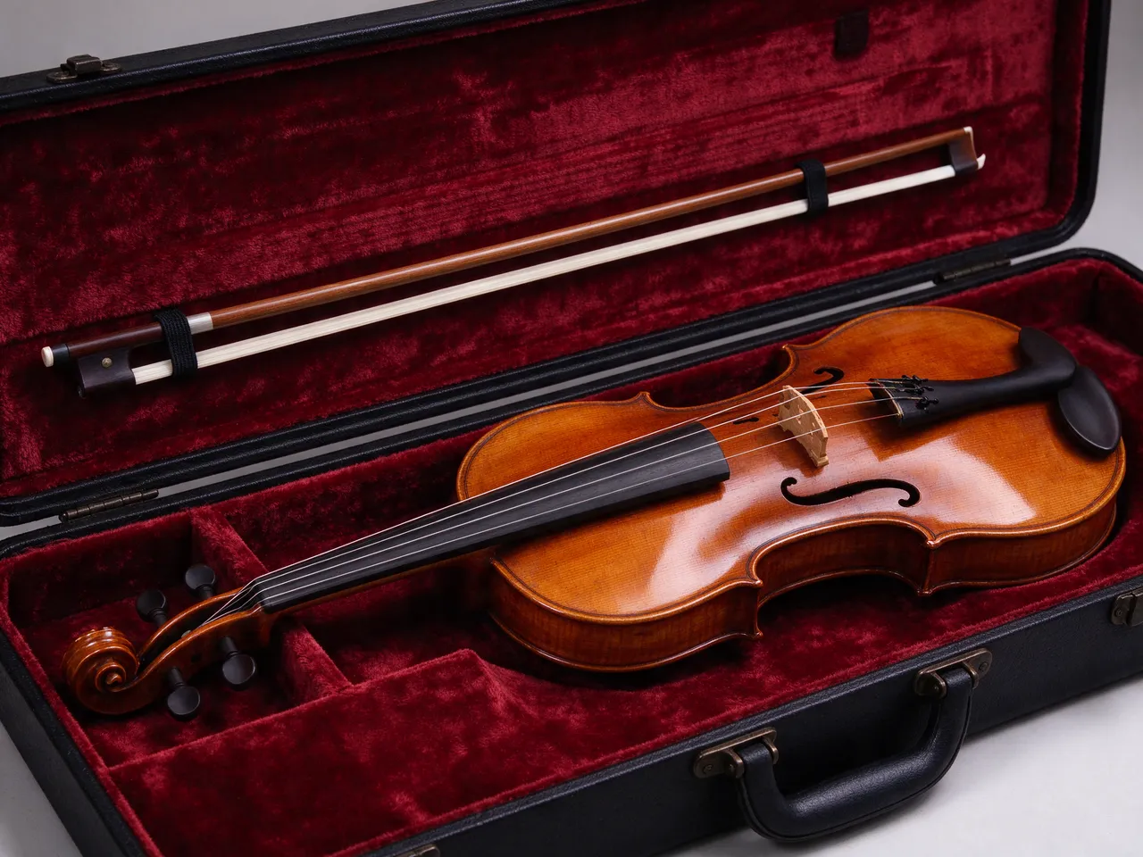 A polished wooden violin rests inside an open hard case lined with dark red velvet, with its bow clipped to the case lid.