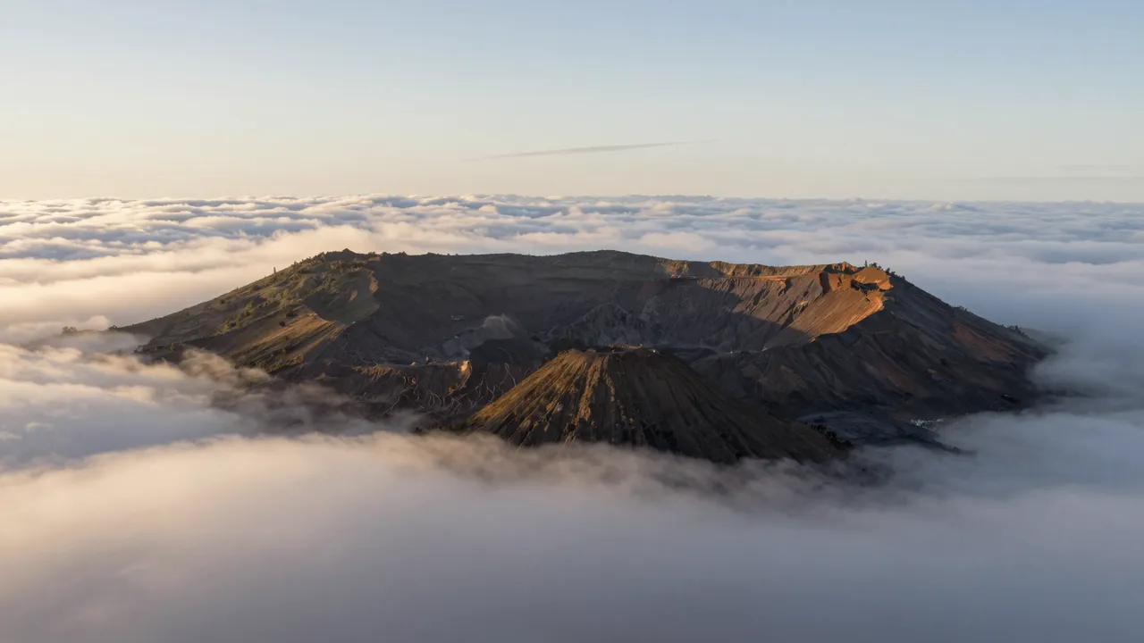 A volcanic crater and cinder cone rise above a sea of soft clouds at warm sunrise