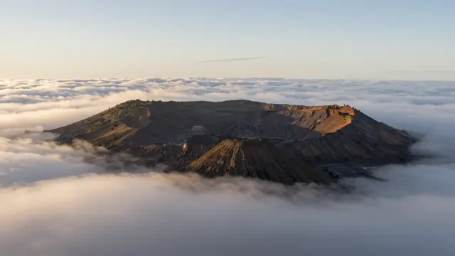 A volcanic crater and cinder cone rise above a sea of soft clouds at warm sunrise