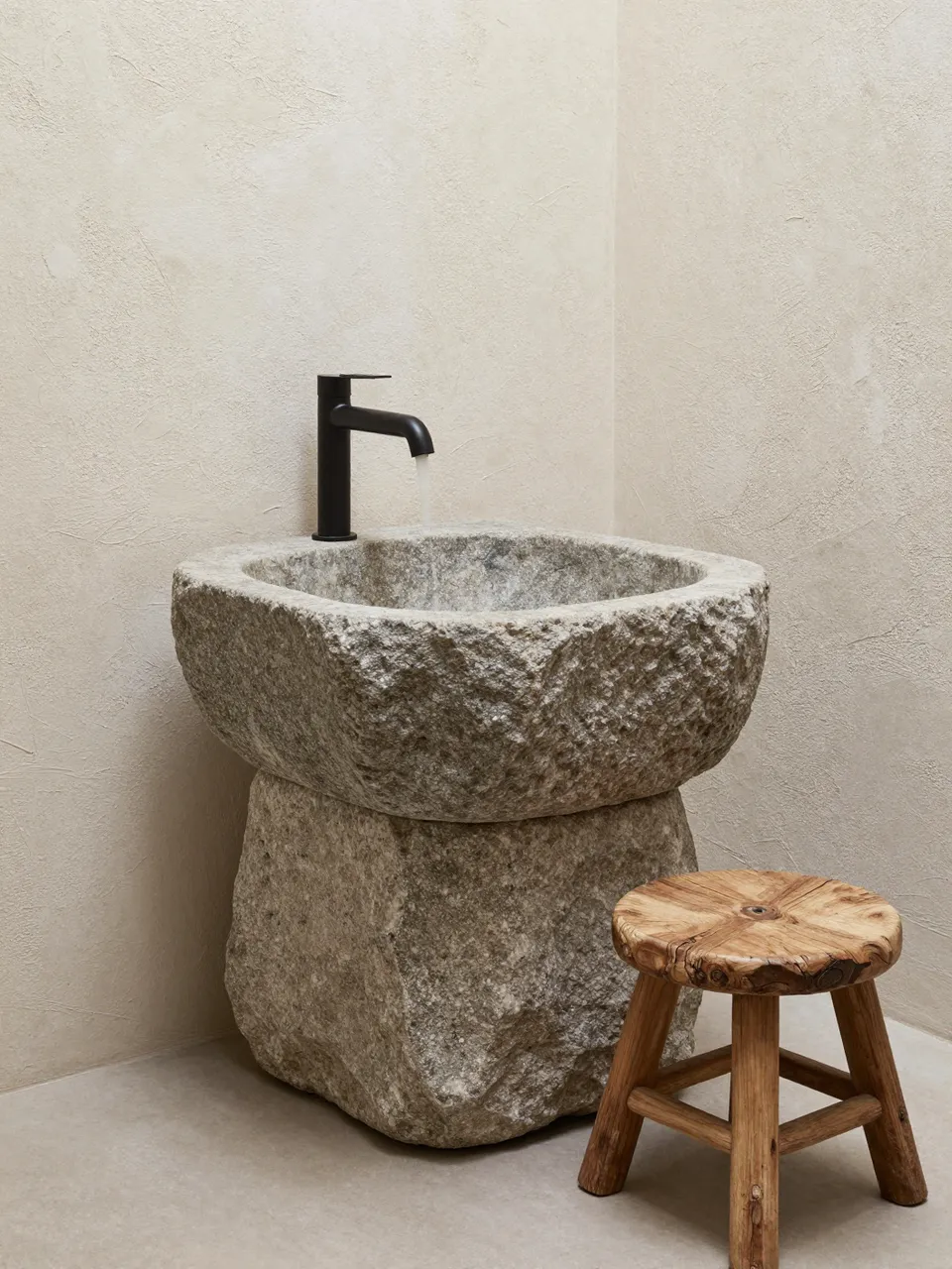 Hand carved stone basin sits next to a weathered wooden stool in a textured plaster bathroom corner