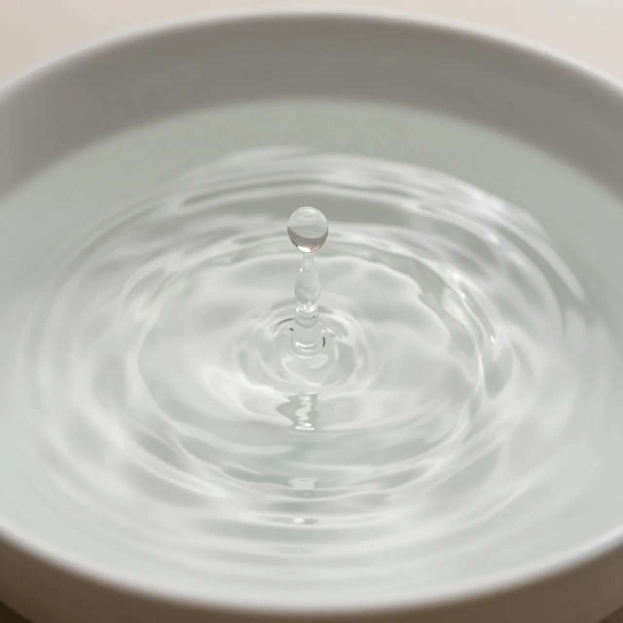 A water drop hovers above ripples spreading across the surface of water in a white bowl.